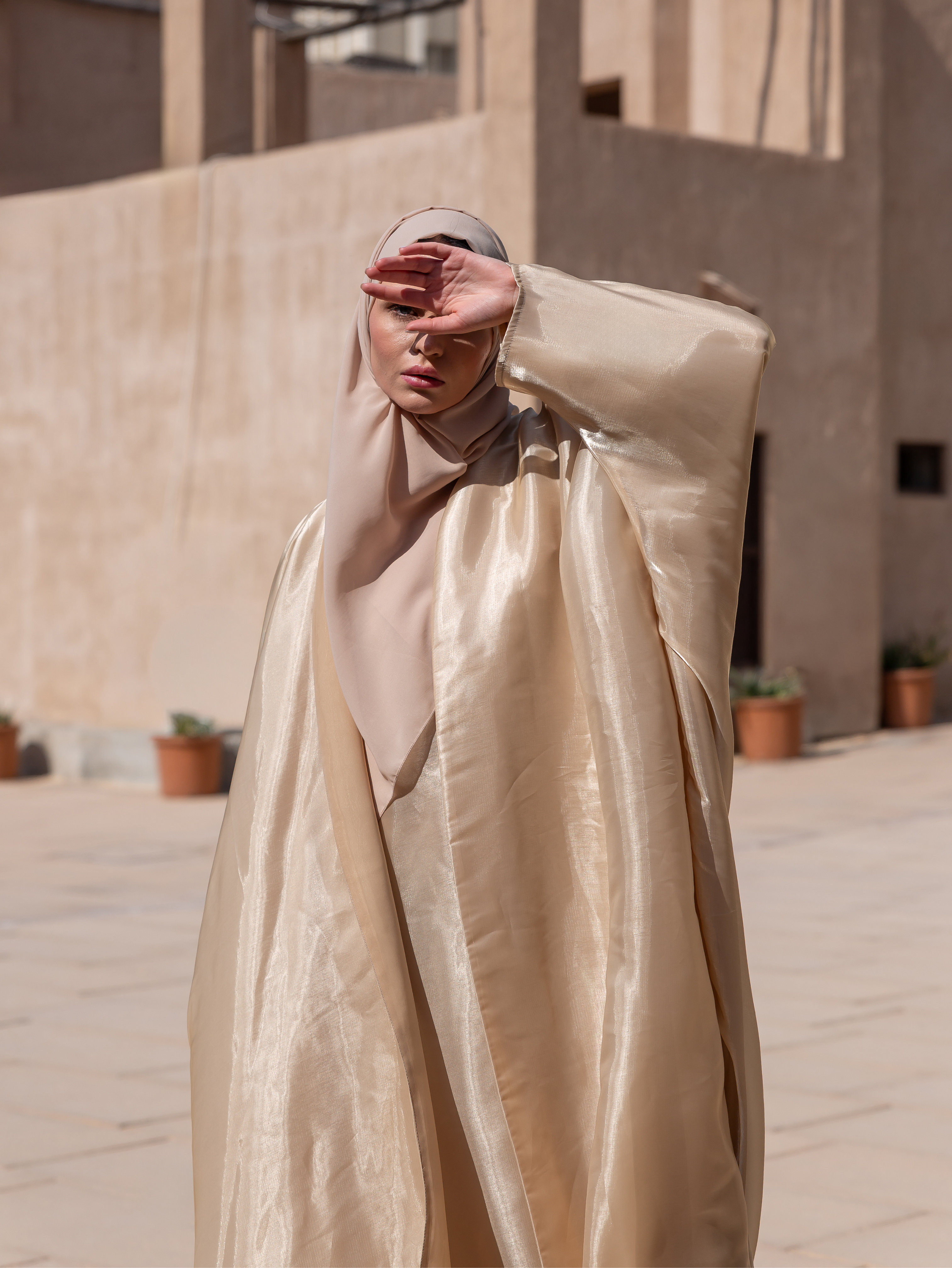 Person wearing a beige hijab and long garment in an outdoor setting with plants and a building.